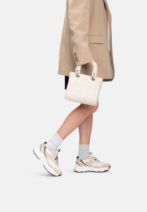 Personne portant des baskets beiges et des chaussettes, tenant un sac matelassé blanc, vêtue d'un manteau beige et d'une jupe noire, marchant sur un fond blanc.