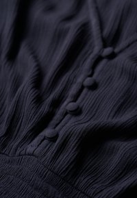 Marineblåt tekstureret stof med vandrette plisseringer og runde, stofbeklædte knapper, der løber ned langs midten. Blødt, strømmende materiale.