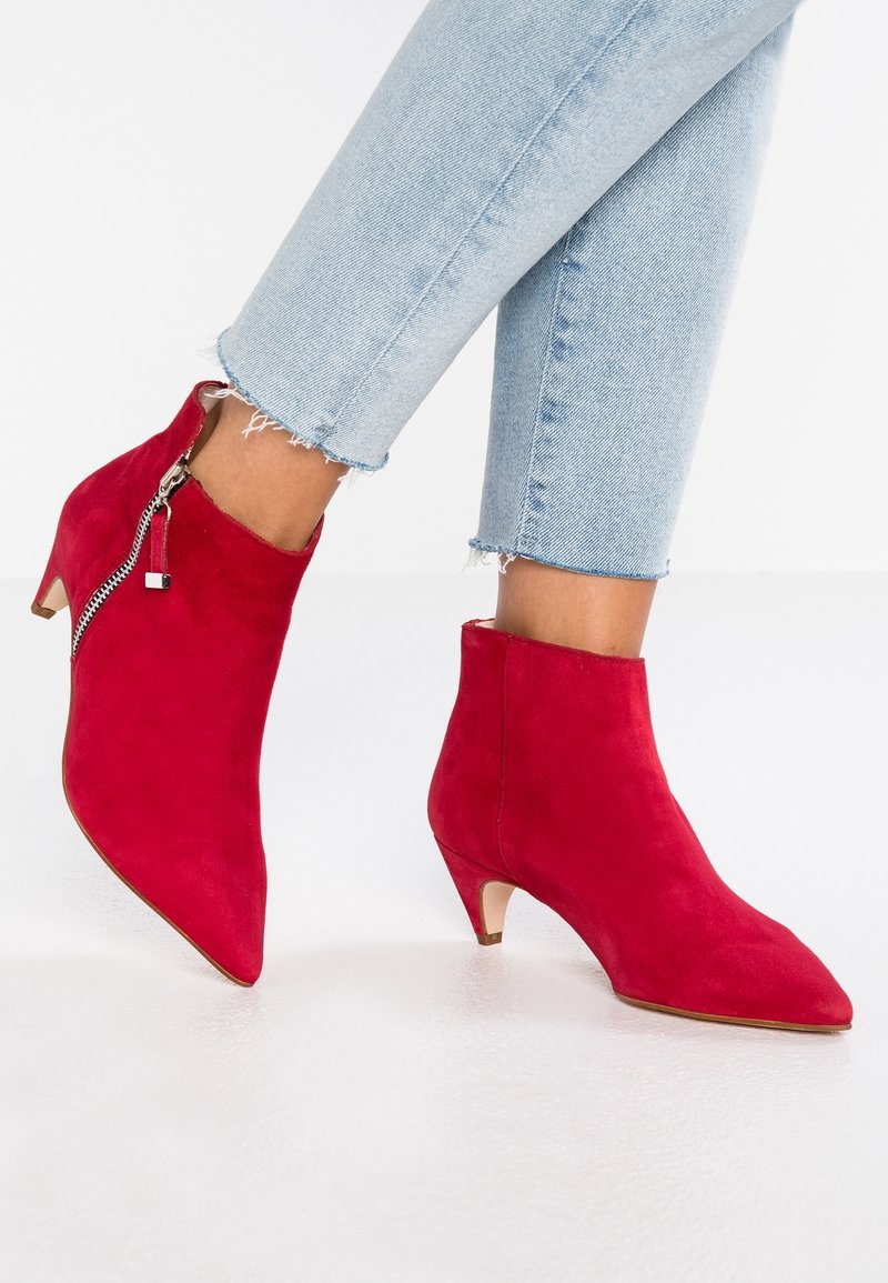 Pieds chaussés de bottines en daim rouge vif avec petits talons et fermetures éclair latérales, associées à un jean bleu clair raccourci à ourlet effiloché.