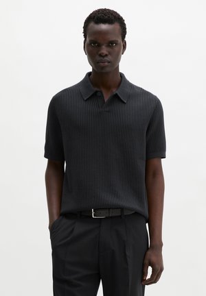 Mężczyzna stojący z rękami w kieszeniach, ubrany w czarną prążkowaną koszulkę polo z krótkim rękawem i czarne spodnie z paskiem.