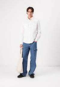 Hvit skjorte med knappestolpe og to brystlommer, sammen med blå vide bukser og svarte sko. Holder en beige tote bag.