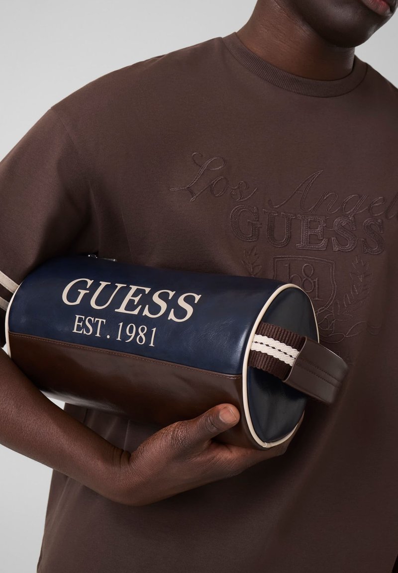 Άτομο που φοράει καφέ μπλούζα Guess κρατάει καφέ και μπλε θήκη Guess με λευκό κείμενο "GUESS EST. 1981" και ριγωτή λαβή.