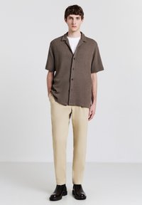 Camicetta marrone a maniche corte con texture, colletto e bottoni neri, abbinata a pantaloni beige e stivaletti neri.