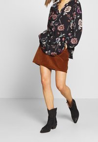 Schwarze Bluse mit floralen Mustern und langen Ärmeln, kombiniert mit einem braunen Cordminirock. Schwarze Stiefeletten runden das Outfit ab.
