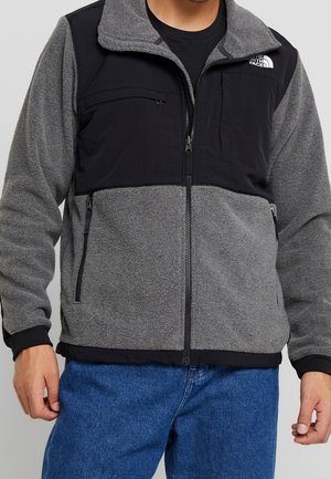 Homme portant une veste polaire zippée North Face grise et noire et un jean bleu, debout devant un fond uni.