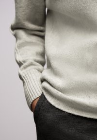 Maglione lavorato a maglia grigio chiaro con polsini a coste, indossato sopra pantaloni scuri e testurizzati. Primo piano che mostra il dettaglio del tessuto e la vestibilità in vita.