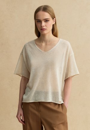 Jonge vrouw met steil blond haar draagt een losse beige V-hals top met korte mouwen en een bruine broek, staand tegen een effen achtergrond.