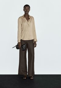 Camicia beige in satin a maniche lunghe con bottoni, abbinata a pantaloni larghi marroni. La modella tiene una pochette nera, in piedi su una superficie scura.