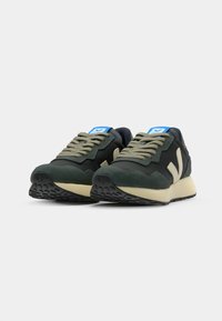 Sport sneakers med mørkegrønt overdel, lys beige midtsåle og svart teksturert yttersåle. Har grå lisser og en blå logoetikett.