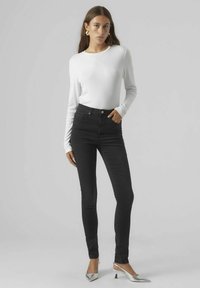 Schwarz-hochgeschnittene Jeans mit schlanker Passform, ausgestattet mit einem Fünf-Taschen-Design und silbernen Beschlägen, kombiniert mit einem langärmeligen weißen Oberteil.