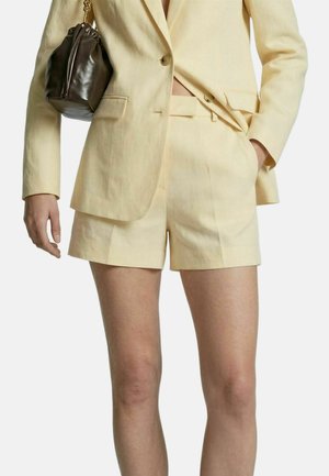 Vrouw draagt een lichtbeige op maat gemaakte blazer en bijpassende korte broek, met een kleine donkerbruine leren handtas over haar schouder.