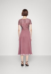 Mauve Kleid mit kurzen Ärmeln, texturiertem Oberteil und fließendem Rock. Durchsichtiger Saum. Trägt dazu schwarze Riemchen-High Heels.
