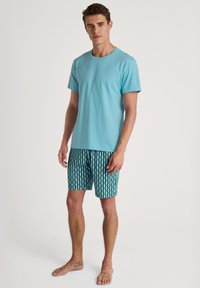 Lichtblauw katoenen T-shirt gecombineerd met patroon shorts in teal met een geometrisch ontwerp, gedragen door een mannelijke model met blote voeten.
