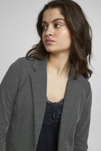 Blazer gris con textura y un sutil patrón de espiga, que presenta un cuello muesca, combinado con una blusa oscura con encaje por debajo.