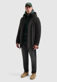 Zwarte gewatteerde jas met capuchon, gedragen over een donkergroene hoodie, gecombineerd met grijze jeans en beige laarzen. Een groene pet maakt de outfit compleet.