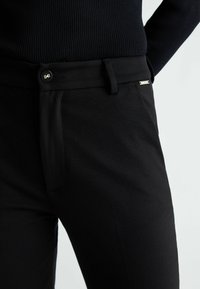 Pantaloni neri in tessuto liscio con un design aderente, dotati di chiusura a bottone e un logo ben visibile vicino alla vita.