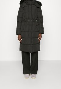 Černý péřový kabát s kapucí lemovanou kožešinou, páskem v oblasti pasu a prošívaným designem. Dlouhé rukávy a dvě přední kapsy. Nosit s širokými kalhotami.