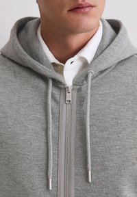 Felpa grigia con zip, dal design aderente, con cordini regolabili e tessuto textured. Indossata sopra una camicia bianca con colletto.
