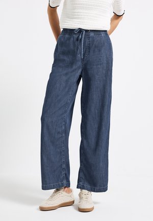 Bredde ben, mørk denim bukser med elastisk talje og snor. Blød tekstur med subtilt slid, parret med beige sneakers.