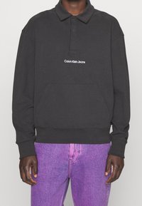 Black, long-sleeve polo sweatshirt with a collar, featuring a front pocket and white "Calvin Klein Jeans" logo. Paired with purple jeans.