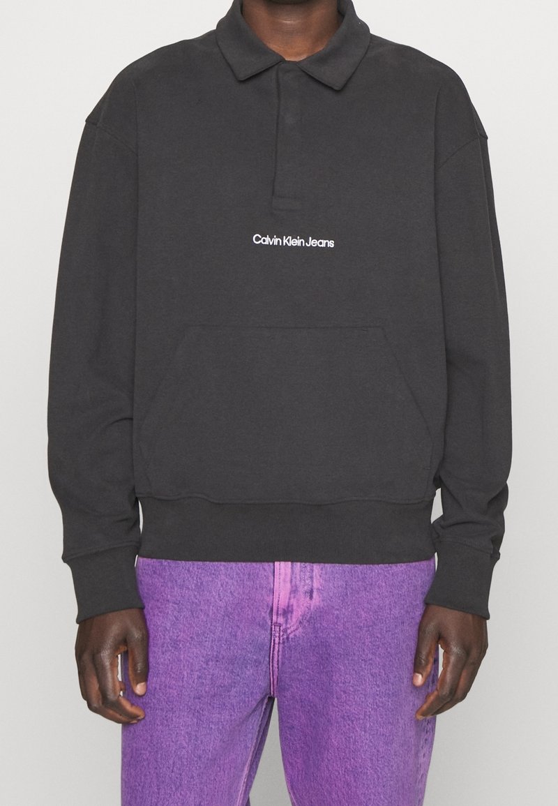 Black, long-sleeve polo sweatshirt with a collar, featuring a front pocket and white "Calvin Klein Jeans" logo. Paired with purple jeans.