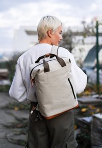 Zaino beige chiaro con accenti marroni, design a risvolto, spallacci regolabili e una tasca frontale con zip. Realizzato in materiale resistente.