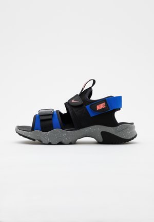 Sandalo sportivo Nike nero e blu con cinturini regolabili, logo rosa e suola grigia maculata, progettato per attività all’aperto.