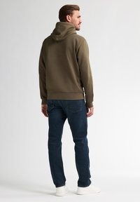 Olivově zelená mikina s kapucí a žebrovaným lemem, spárována s tmavě modrými džíny a bílými teniskami, zobrazená zezadu.