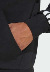 Sweatshirt preto com bolso frontal e punhos canelados. Apresenta um tecido texturizado e detalhes em listras brancas nas mangas.