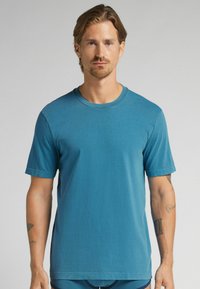 Muž se světle hnědými vlasy a vousy, oblečený v jednoduchém modrém tričku s kulatým výstřihem, stojící na neutrálním pozadí.