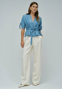 Model in einer hellblauen Wickelbluse mit gebundenem Bund, kombiniert mit weißen, weiten Hosen und spitzen, weißen Schuhen, vor einem neutralen Hintergrund.