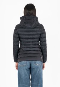 Zwarte pufferjas met horizontale quilt, aangehechte capuchon en elastische manchetten. Draag het over een blauwe spijkerbroek, met een logo op de onderste zoom.