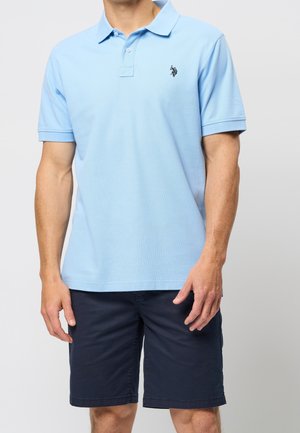Man draagt lichtblauw poloshirt met korte mouwen en klein zwart logo en marineblauwe knielange korte broek tegen een effen achtergrond.