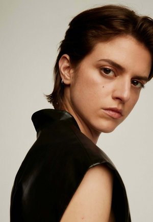 Top de cuero negro con cuello alto y diseño sin mangas. El modelo tiene el cabello corto y lleva un pendiente de aro, mirando ligeramente hacia la izquierda.