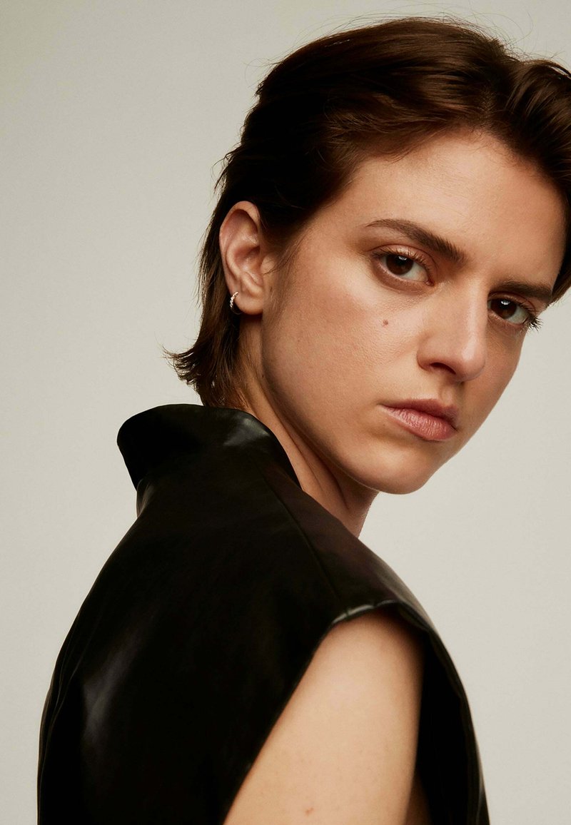 Top de cuero negro con cuello alto y diseño sin mangas. El modelo tiene el cabello corto y lleva un pendiente de aro, mirando ligeramente hacia la izquierda.
