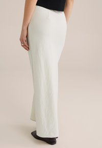 Bijela, naborana suknja s ravnim siluetom, srednje visokim strukom i detaljem na bočnom šavu, uparena s crnim cipelama do gležnjeva.