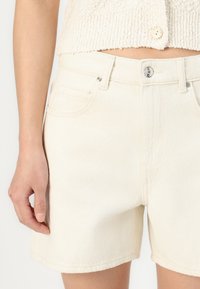 Světle béžové denimové šortky s klasickým střihem, které mají přední kapsy, zapínání na knoflík a jemnou texturu. Spodní lem končí nad kolenem.