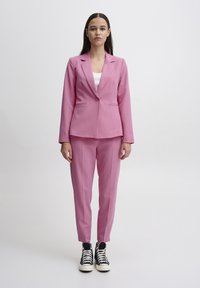 Růžový blazer s jedním knoflíkem a klopovými límci, v kombinaci s odpovídajícími zúženými kalhotami. Pod ním bílý top a černé vysoké tenisky.