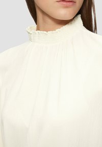 Blouse blanche avec un col haut et volanté. Comprend un tissu doux et texturé avec des plis sur le devant, créant une silhouette fluide.