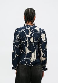 Bluza cu model floral în albastru marin și crem, cu mâneci lungi, țesătură texturată, guler cu volane și manșete elastice. Detaliul de la spate include o cusătură.