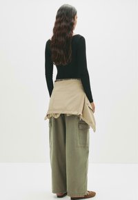 Svart långärmad topp, lösa olivgröna cargo-byxor, beige jacka knuten runt midjan, slät textur, monokrom färgskala, avslappnad stil.