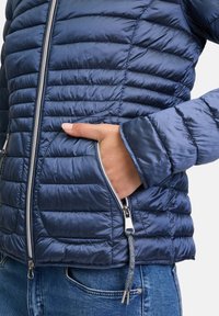 Marineblauw geïsoleerde jack met een geborduurd patroon, een ritssluiting aan de voorkant en zijzakken. De stof heeft een gladde glans; elastische manchetten zorgen voor een snug fit.