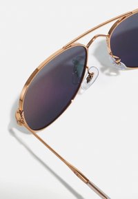Ochelari de soare aviator cu nuanță aurie, lentile în degrade întunecat, pernă de nas transparentă și un design de cadru elegant și unghiular. Accente argintii lângă balamale.