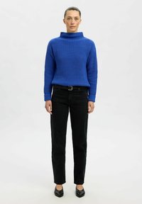Blauer gerippter Rollkragenpullover, lange Ärmel, kombiniert mit schwarzen hochgeschnittenen Jeans und schwarzen spitz zulaufenden Ballerinas. Einfarbigem Hintergrund.