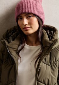 Jongeman met een roze gebreide beanie, een witte shirt en een olijfgroene gewatteerde jas tegen een neutrale achtergrond.