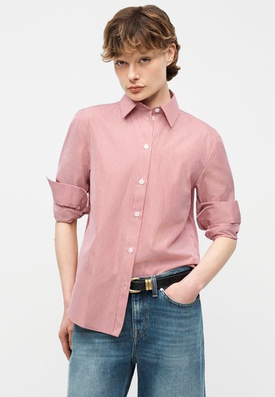 Νεαρό άτομο με κοντά μαλλιά, ντυμένο με πουκάμισο με κόκκινες και λευκές κάθετες ρίγες, με κουμπιά και μανίκια γυρισμένα, και μπλε τζιν, με το χέρι στην τσέπη.