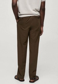 Braune Hosen aus glattem Stoff, mit schmaler Passform, Gesäßtaschen und klassischem Schnitt, kombiniert mit schwarzen Sandalen.