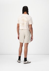 Tricou din bumbac bej deschis cu un imprimeu grafic pe spate, asociat cu pantaloni scurți texturat gri deschis. Tenis și șosete negre completează look-ul.