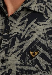Košulja na dugih rukava s tamnozelenim i crnim apstraktnim uzorkom. Uključuje zlatni izvezeni logotip krila i crne gumbe. Tekstura tkanine.