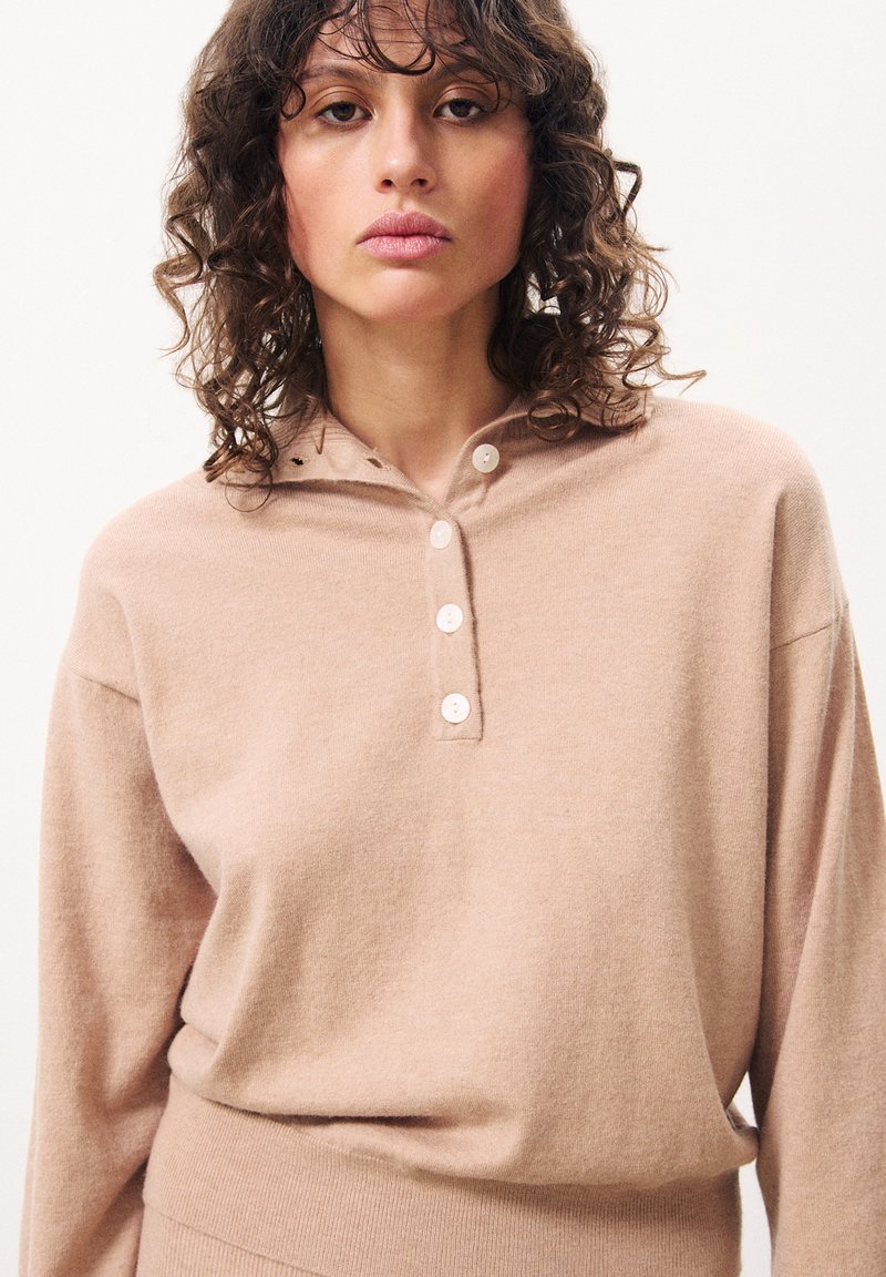 Lichtbruine gebreide pullover met een capuchon, voorzien van een drieknoopsluiting. Zachte textuur en een relaxed fit, met subtiele ribbelstructuur aan de onderkant.